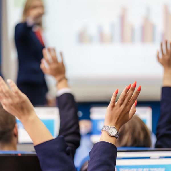 Students raising hands to answer or ask question while attending classes at modern classroom at university. Blurred professor picking student up.